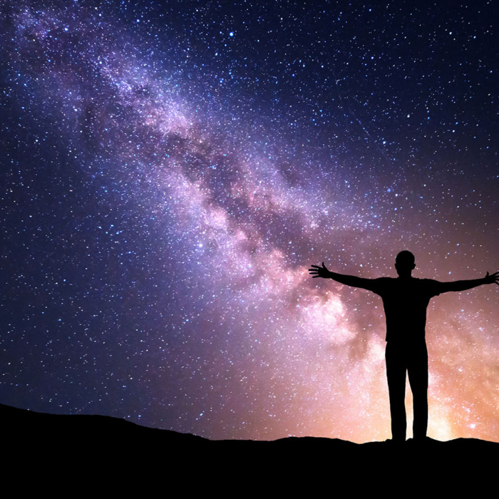 Night colorful landscape with Milky Way, yellow city lights and silhouette of a standing sporty man with raised up arms on the mountain. Beautiful Universe. Travel background with purple sky full of stars