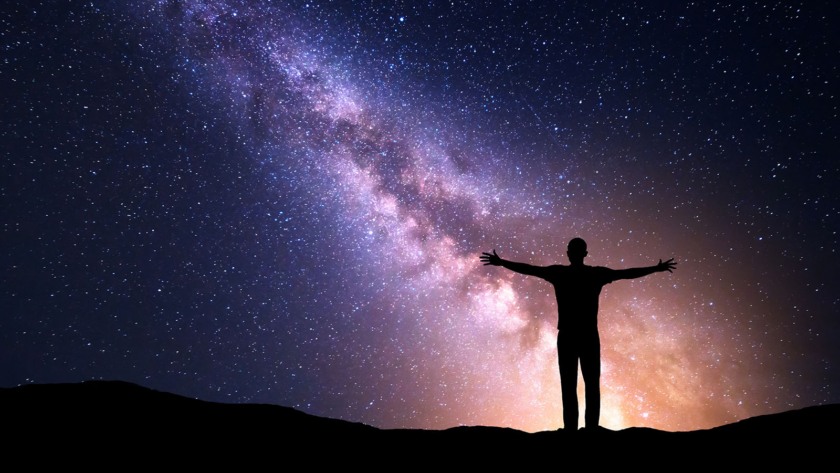 Night colorful landscape with Milky Way, yellow city lights and silhouette of a standing sporty man with raised up arms on the mountain. Beautiful Universe. Travel background with purple sky full of stars